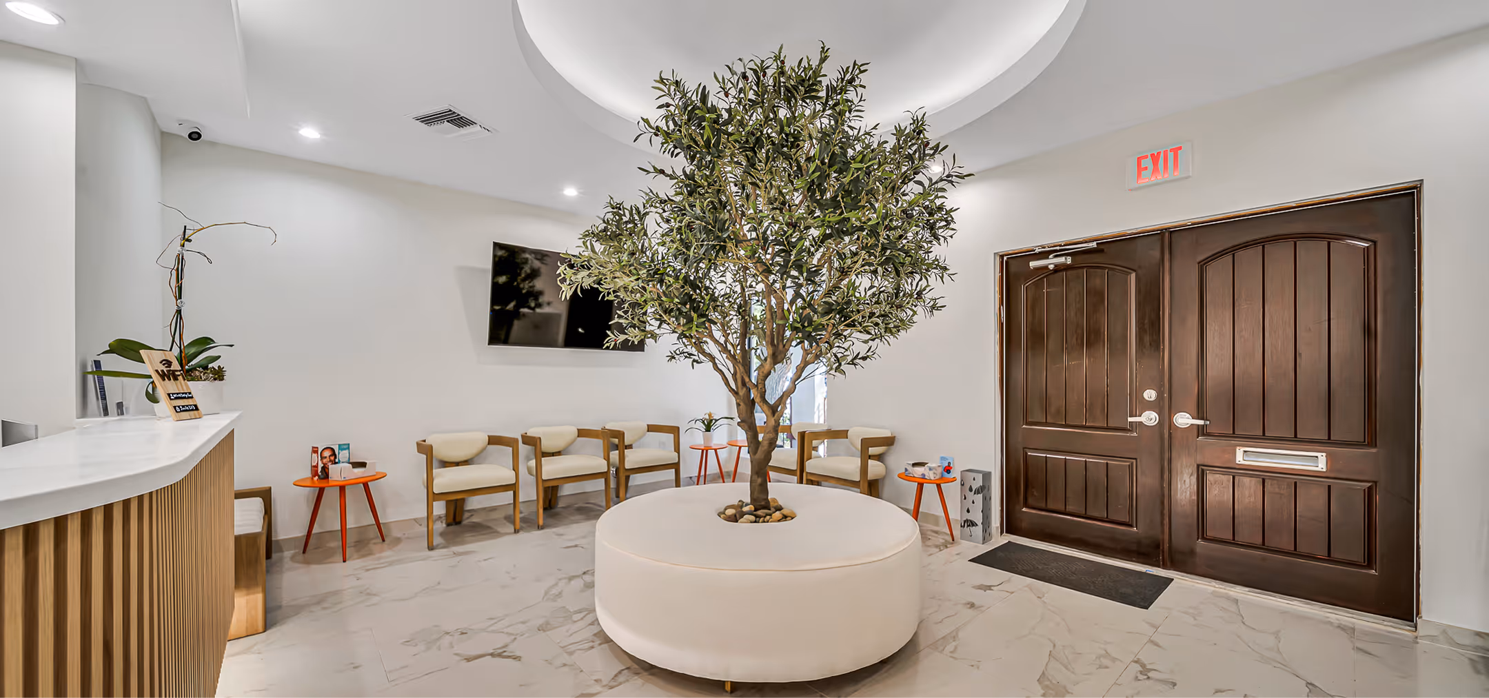 Modern waiting area with round white seating around a central leafy indoor tree, wooden chairs, small orange tables, and a dark double door under an exit sign.