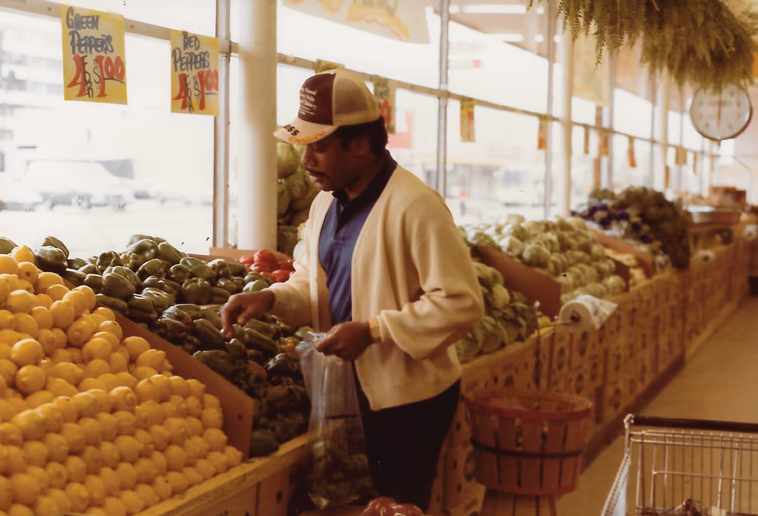 Man wearing a cardigan and baseball cap selecting green bell peppers at an indoor produce market with lemons and other vegetables nearby.