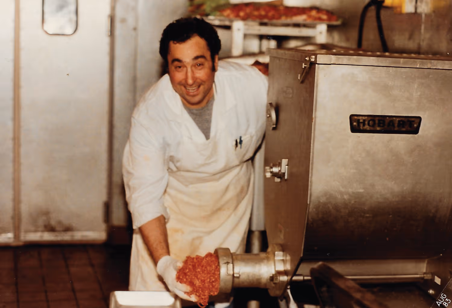 Man in white apron and gloves smiling while operating a Hobart meat grinder with ground meat coming out in a kitchen setting.
