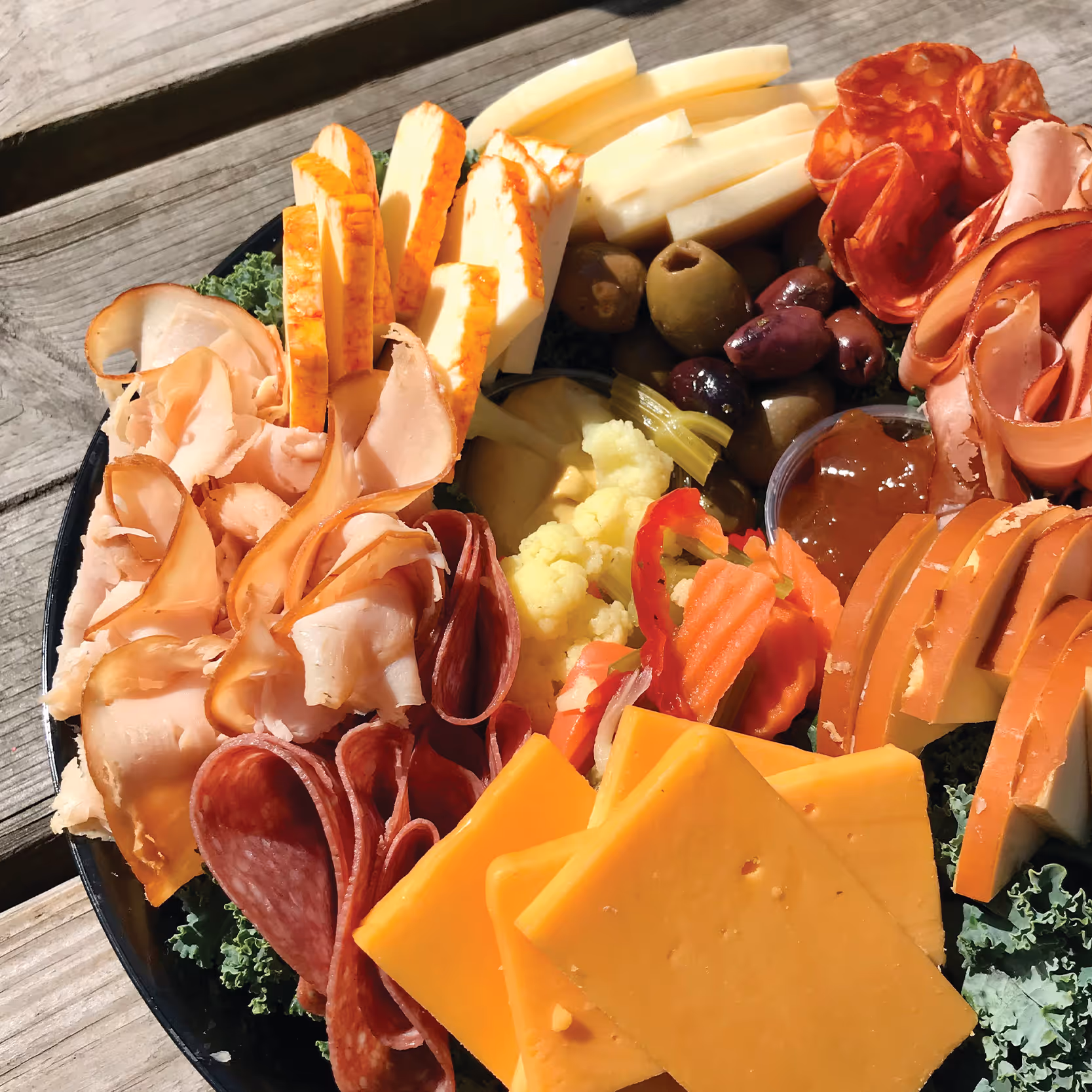 Meat and cheese board with sliced cheddar, provolone, smoked turkey, salami, olives, pickled vegetables, and a dipping sauce on kale.
