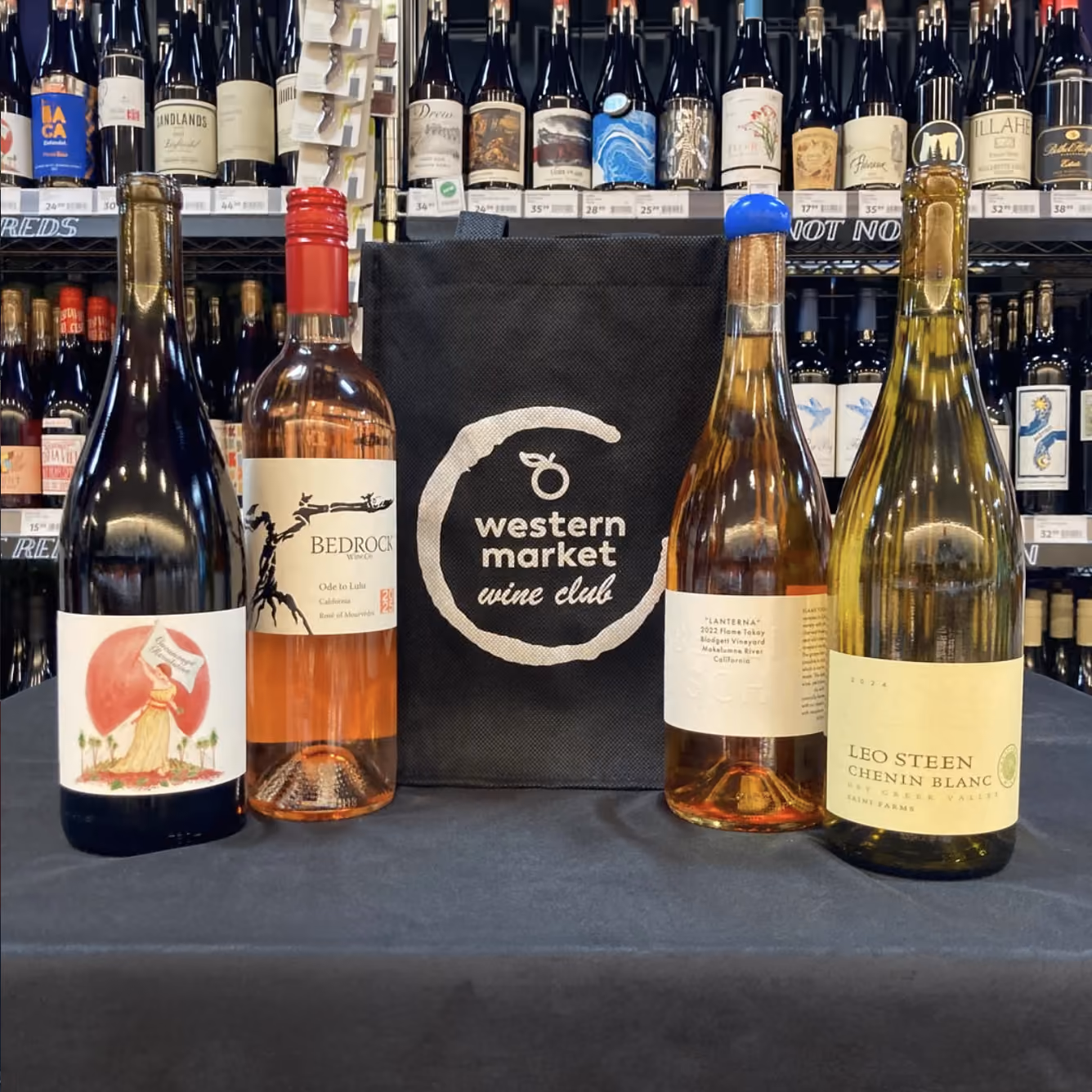 Four wine bottles displayed with a black bag reading 'western market wine club' on a black cloth table.