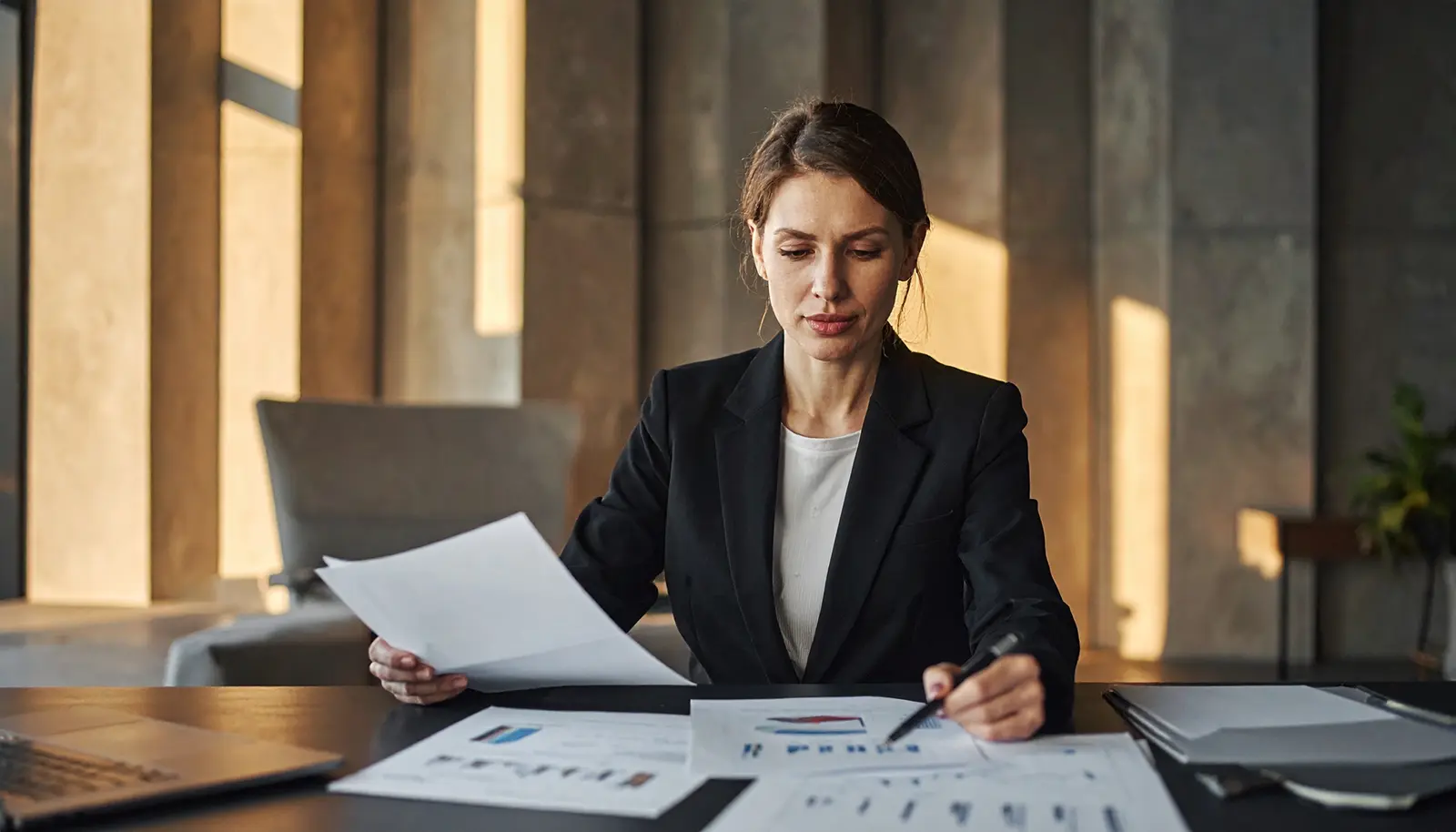 Professional woman reviewing financial documents at a desk, focused downward in concentration, warm side lighting.