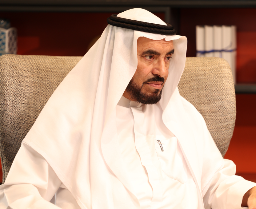 Middle-aged Arab man wearing traditional white kandura and headscarf sitting on a textured chair with bookshelves in the background.