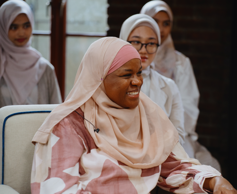 Smiling woman wearing a beige hijab and patterned dress seated, with three women in hijabs in the background.