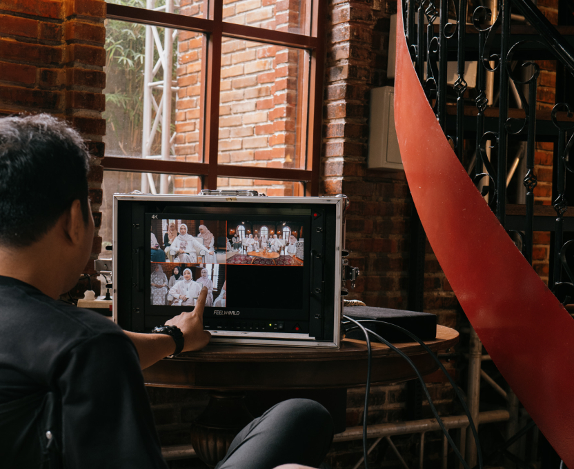 Person pointing at a multi-camera video feed monitor showing a group of women in white outfits inside a brick-walled room.