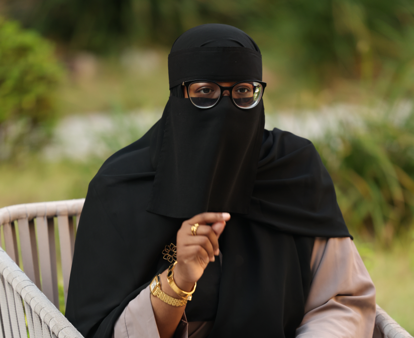 Woman wearing a black niqab and glasses sitting outdoors on a chair with greenery in the background.