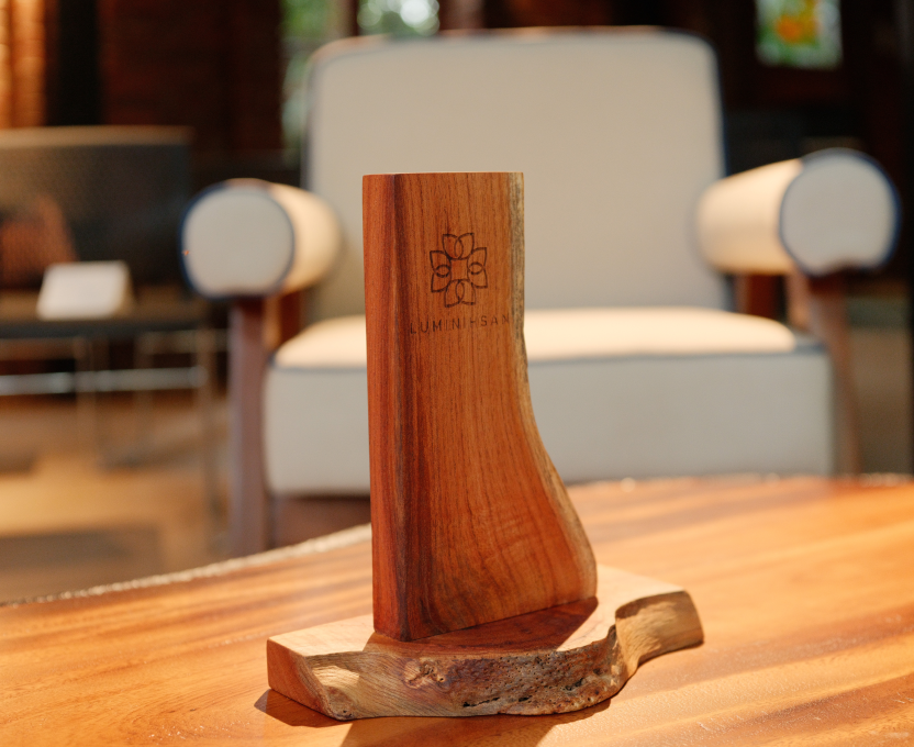 Wooden decorative stand with the engraved text 'LUMINIHISAN' and a flower-like logo, placed on a wooden table with a blurred cushioned armchair in the background.