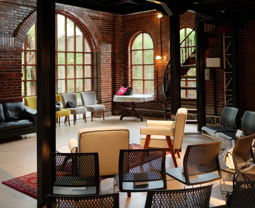 Cozy room with exposed brick walls and large arched windows, furnished with various chairs and a round table with bags on it.