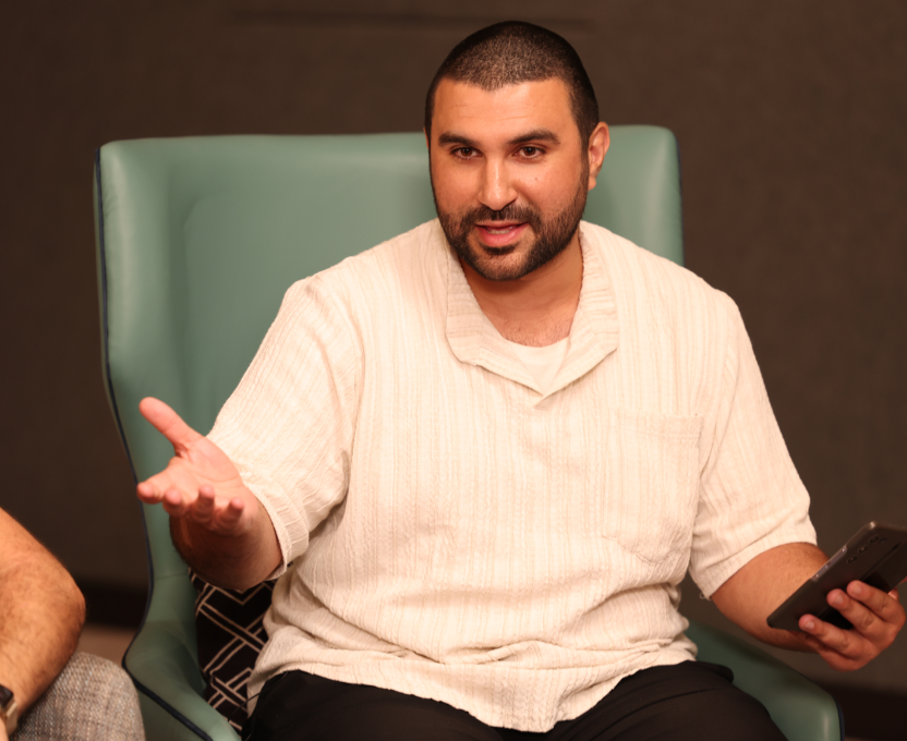 Man with short hair and beard sitting on a teal chair, gesturing with one hand and holding a phone in the other.