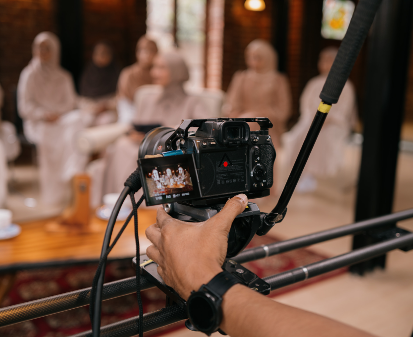 Hand operating a video camera on a tripod capturing a group of women sitting indoors, some wearing hijabs.