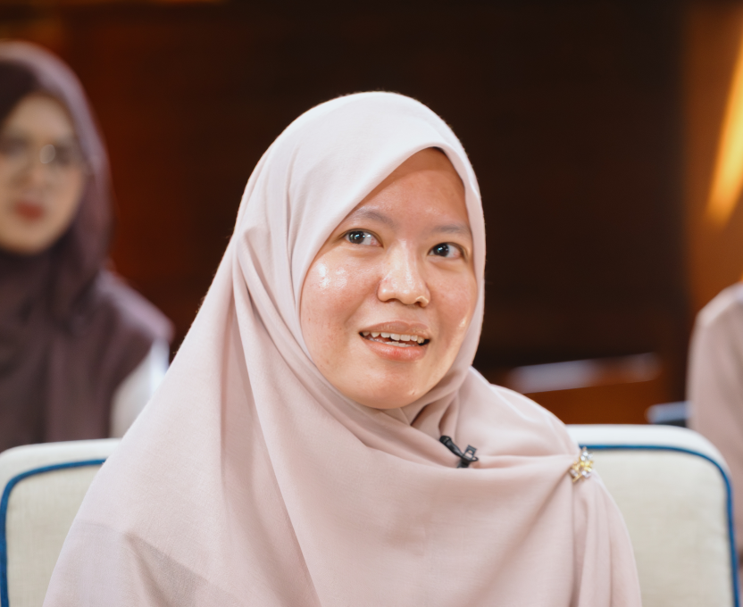 Woman wearing a pale pink hijab speaking in a well-lit room with blurred people in the background.