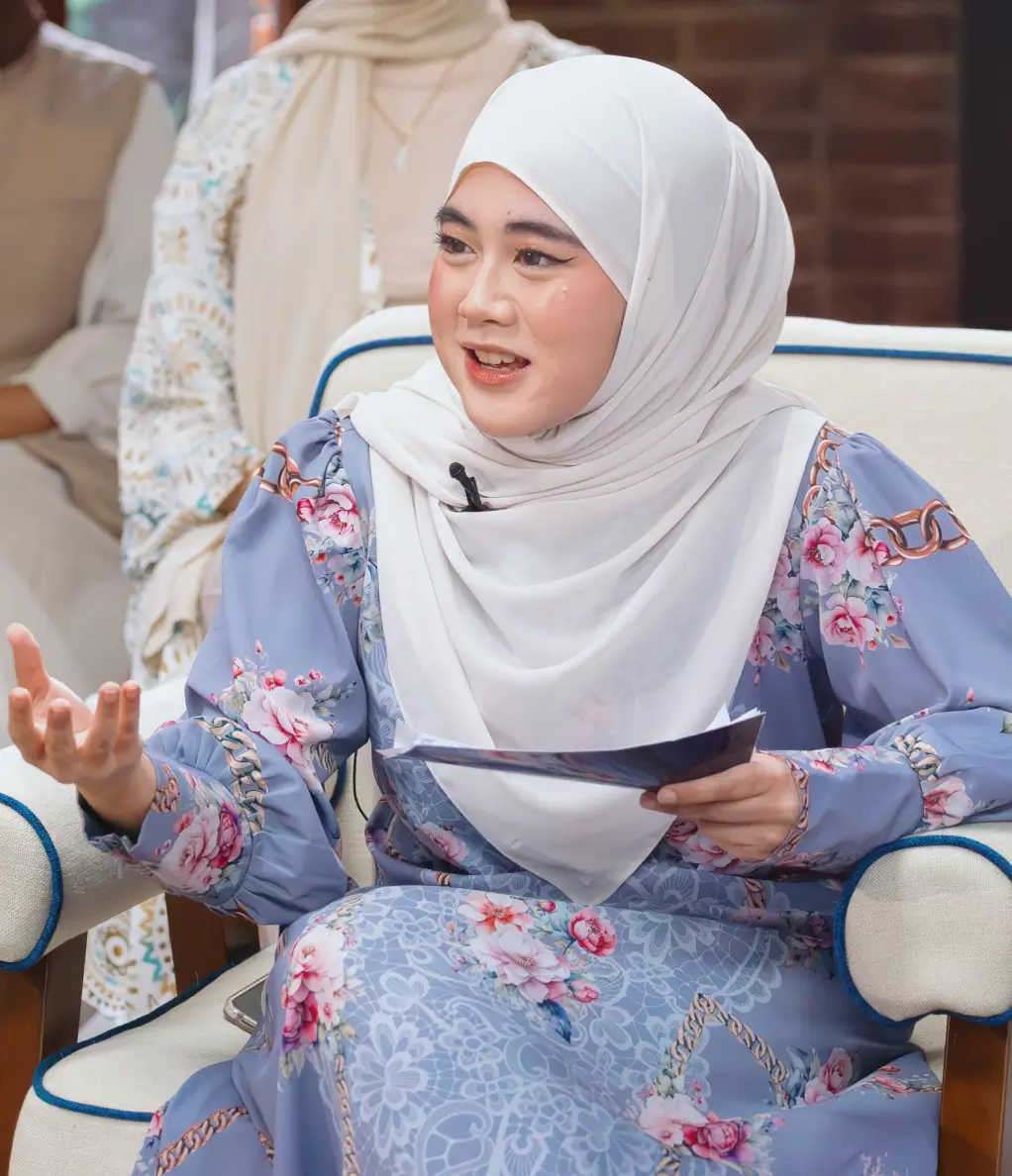 Woman in a white hijab and floral-patterned blue dress speaking while seated on a beige chair with blue piping.