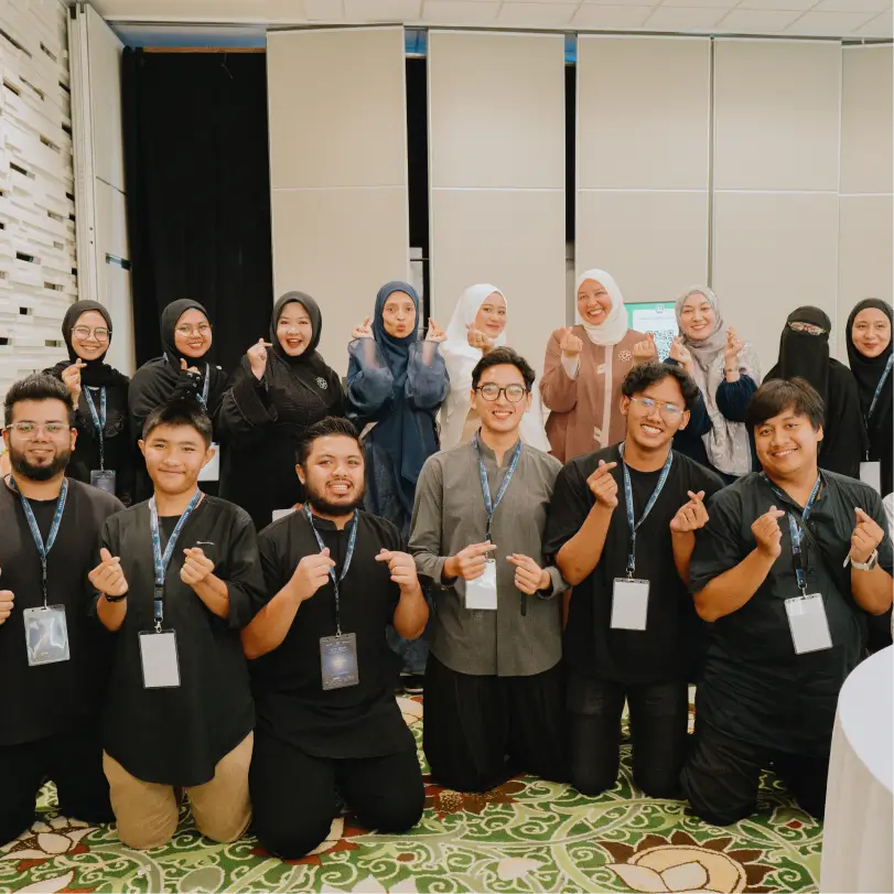 Group of diverse young adults, both men and women wearing hijabs, posing indoors on a patterned carpet, making finger heart gestures and smiling.