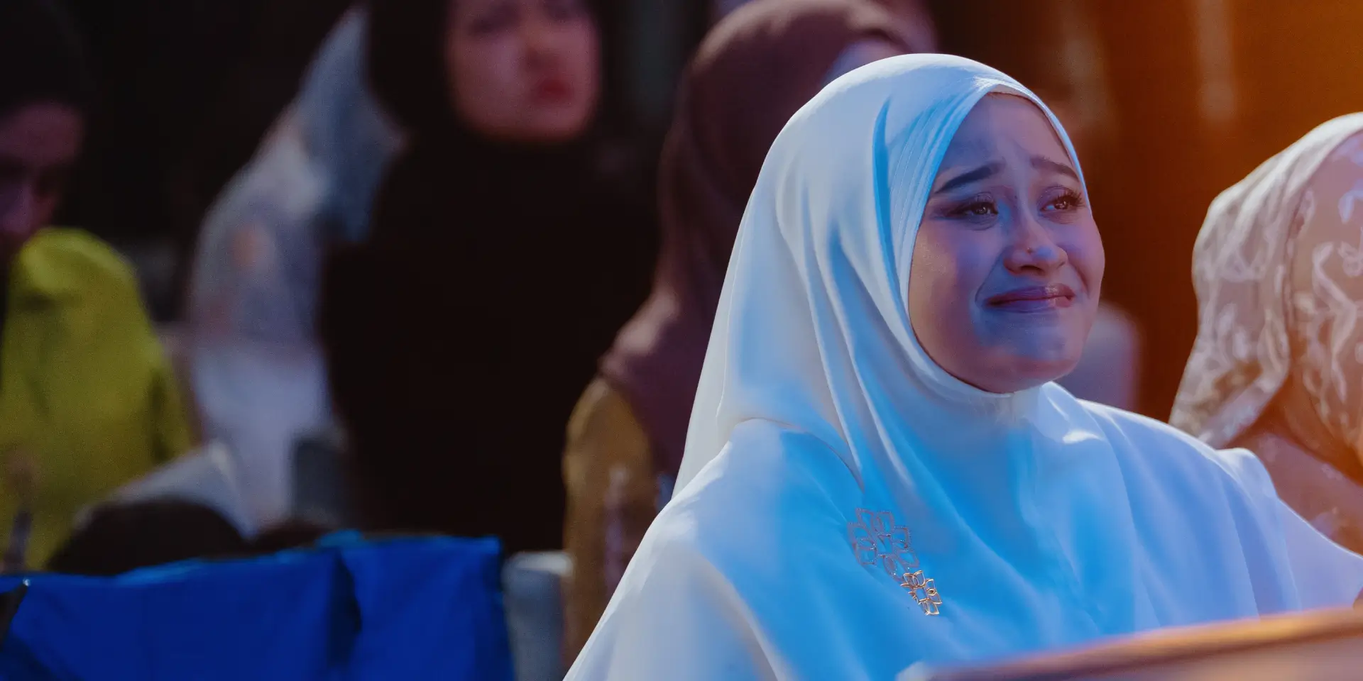 Woman in white hijab looking emotional among other women in headscarves in a dimly lit setting.