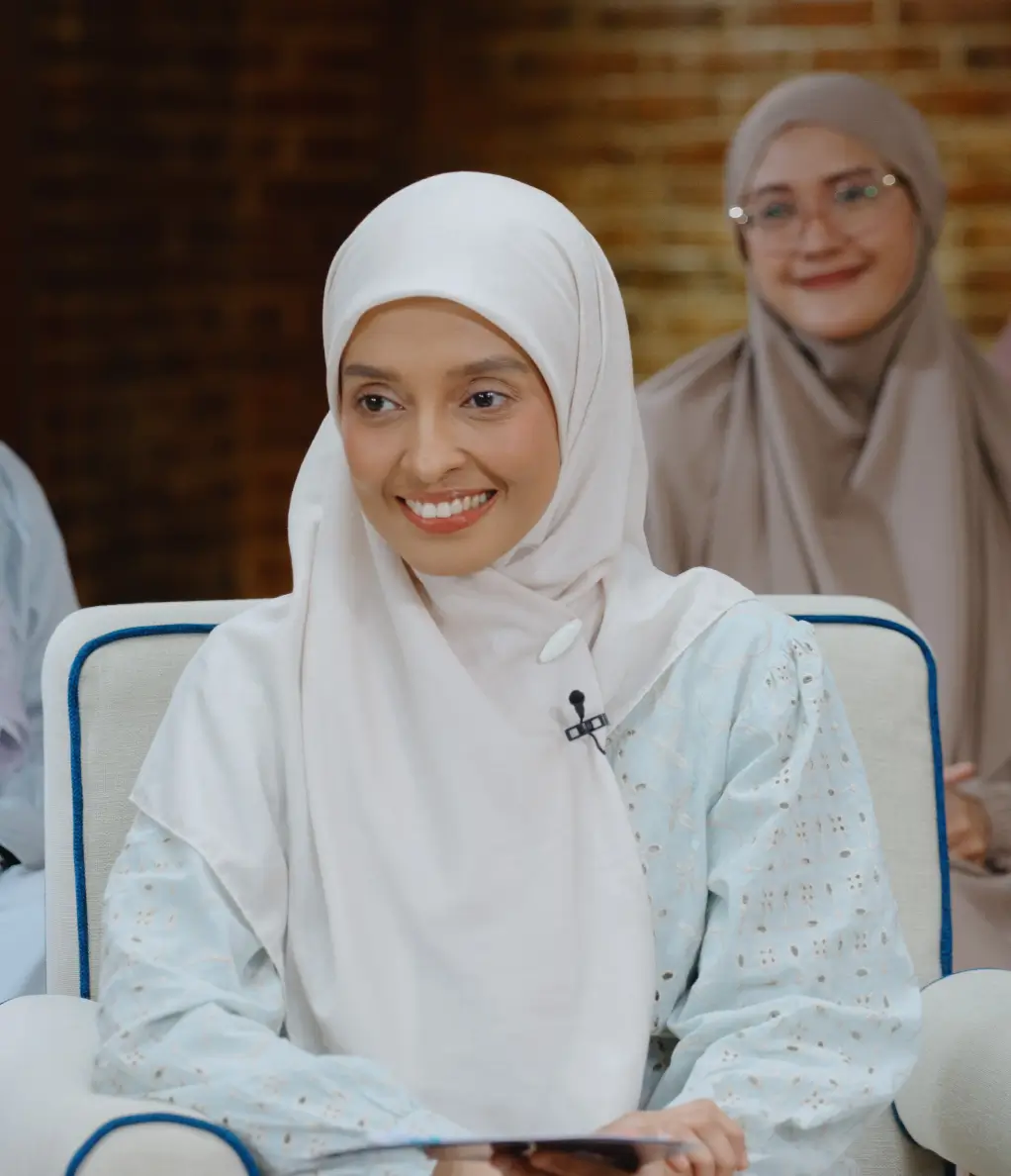 Smiling woman in a white hijab and light blue patterned top sitting on a chair, with another woman in a beige hijab blurred in the background.