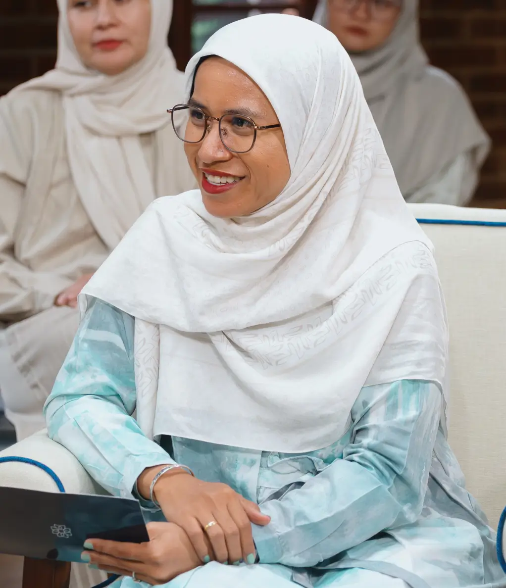Smiling woman wearing glasses and a white hijab seated and holding a script card, with two other women in hijabs blurred in the background.