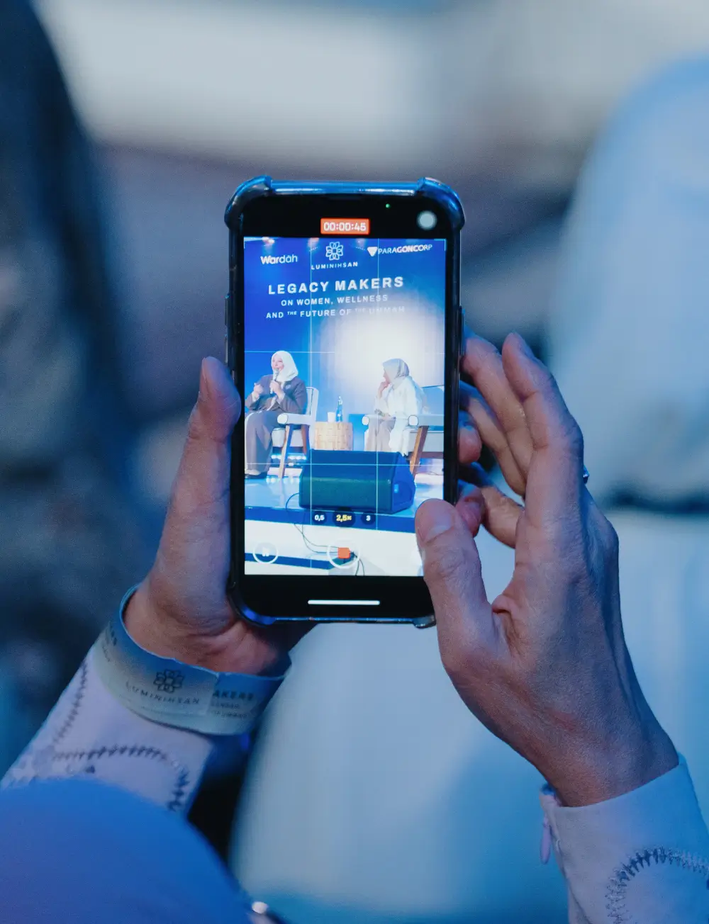 Hands holding a phone recording a panel discussion titled 'Legacy Makers on Women, Wellness, and the Future'.