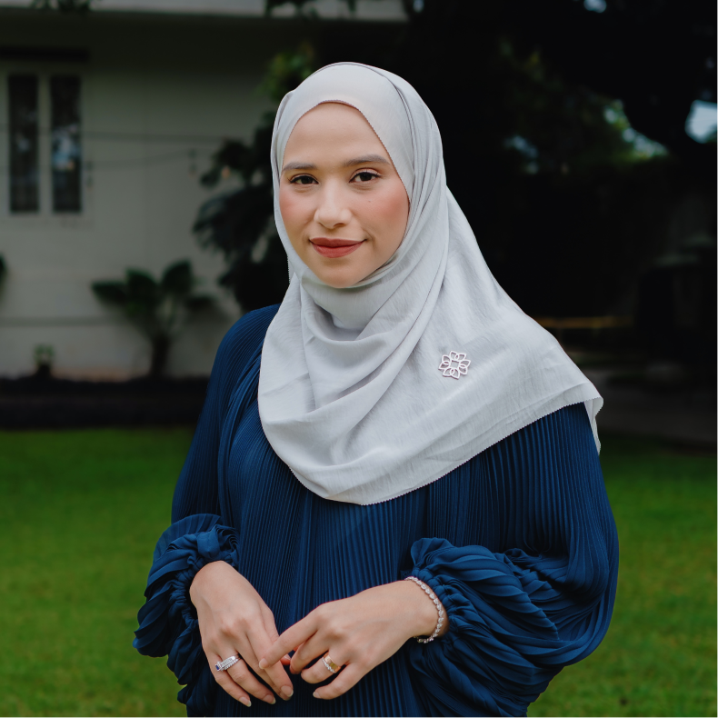 Woman wearing a light gray hijab and a navy blue pleated dress standing outdoors on green grass.