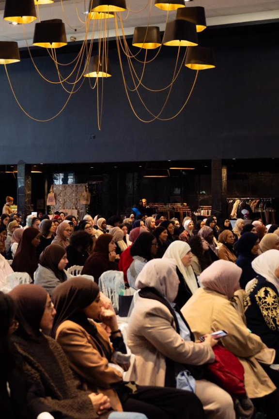 Large group of women wearing hijabs seated attentively in a dimly lit conference room with modern hanging lamps.