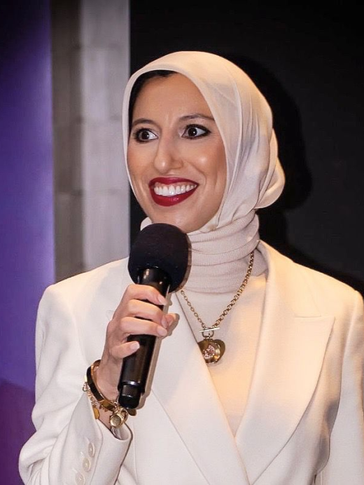 Woman wearing a white hijab and blazer, holding a microphone, and smiling while speaking.