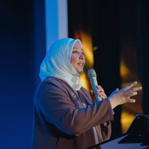 Smiling woman in a white hijab and brown dress speaking into a microphone at a podium in a dimly lit room.