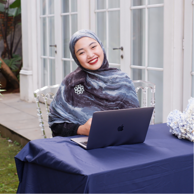 Smiling woman wearing a patterned hijab sitting at a table with a laptop outdoors in front of glass-paneled doors.