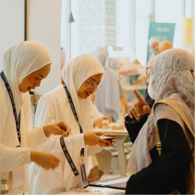 Two women in white hijabs wearing event lanyards assist a visitor in a headscarf at a registration or information desk indoors.