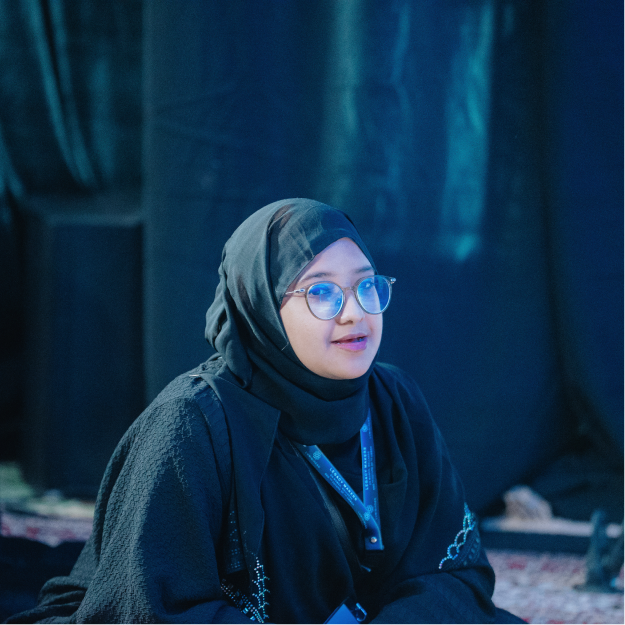 Woman wearing glasses and a black hijab sitting indoors against a dark blue background.