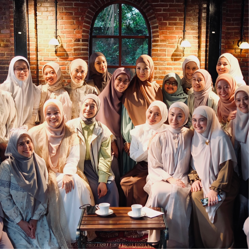 Group of women wearing hijabs posing and smiling together inside a cozy room with brick walls and a window showing greenery outside.