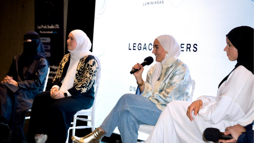 Four women wearing hijabs seated in a panel discussion, with one woman speaking into a microphone.