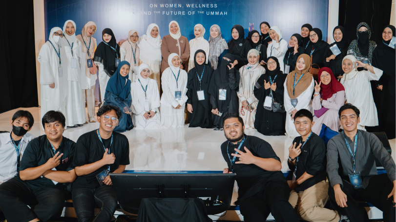 Group photo of diverse women wearing hijabs and men seated in front, posing together on a stage with a backdrop about women, wellness, and the future of the ummah.
