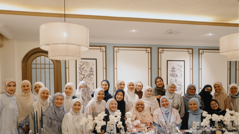 Group of women wearing hijabs, smiling and posing in a warmly lit elegant room with floral decorations on the table.