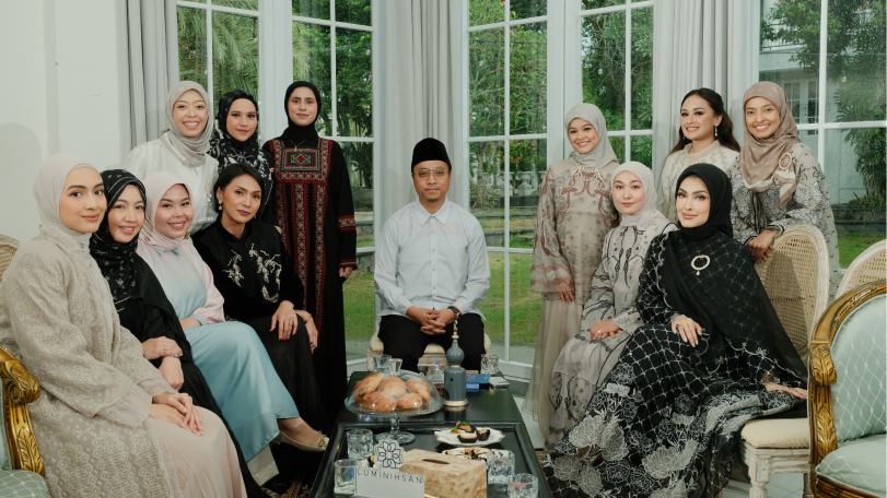 Group of people dressed in traditional attire sitting and standing around a coffee table inside a well-lit room with large glass windows.