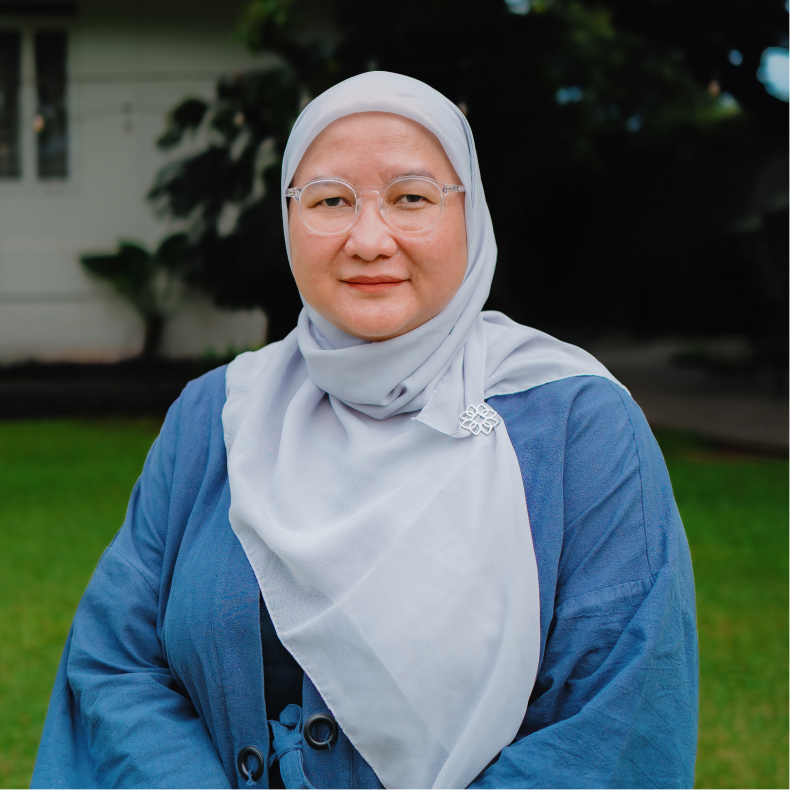 Woman wearing eyeglasses and a light gray hijab with a blue outer garment, posing outdoors with a blurred green lawn and house in the background.
