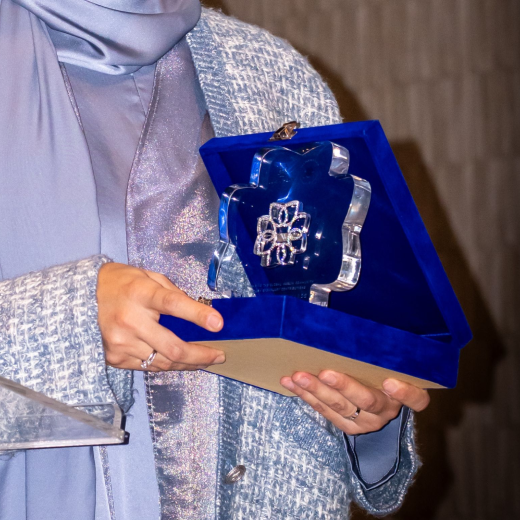 Person wearing a light blue hijab and textured coat holding an open blue velvet box with a luminihsan logo glass award inside.