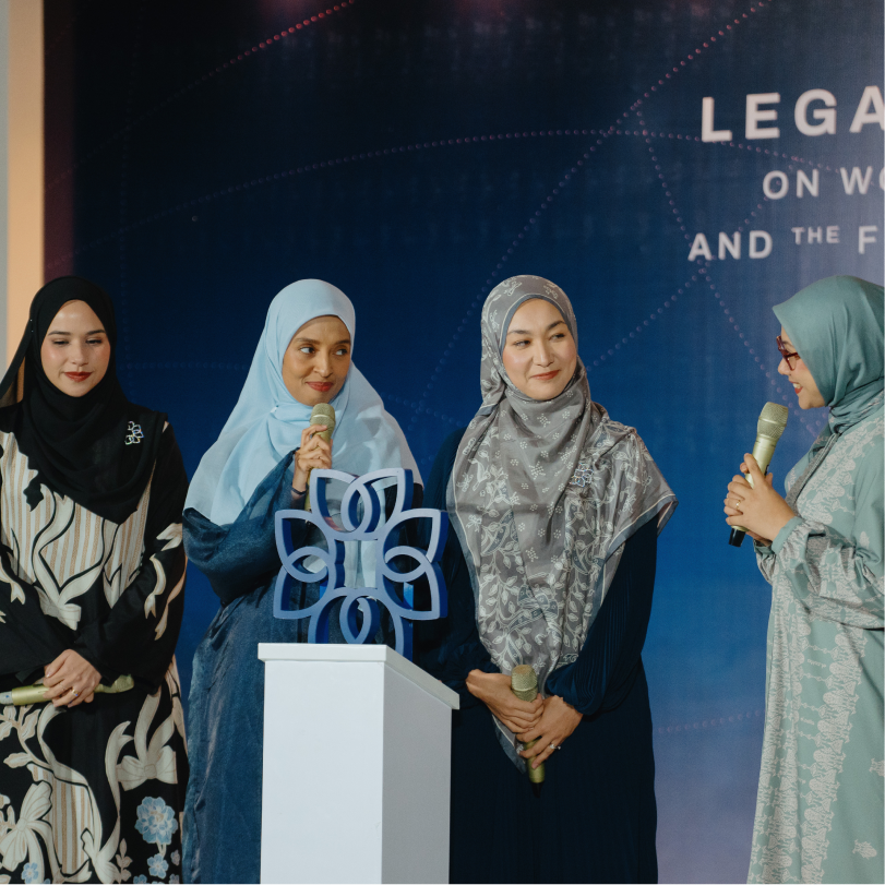 Four women wearing hijabs stand on a stage, holding microphones, with a blue luminihsan logo emblem on a white pedestal in front of them.
