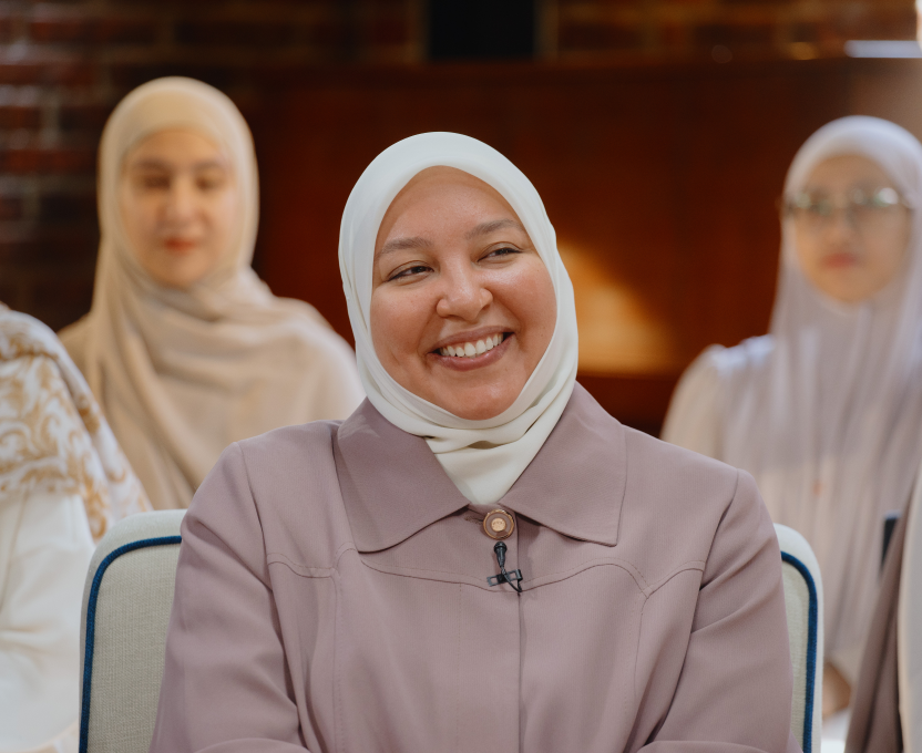 Smiling woman wearing a white hijab and lavender dress, with two other women in hijabs blurred in the background.