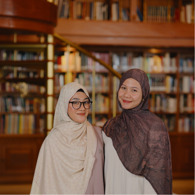 Two women in hijabs smiling in front of a wooden bookshelf filled with books inside a library.