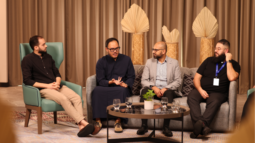 Four men seated in a casual discussion setting with a round table holding water glasses and a small plant in front of them.