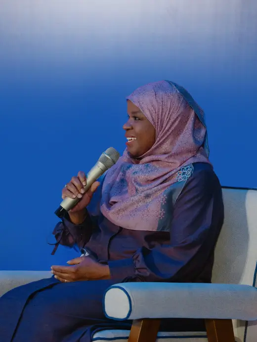 Woman wearing a lilac hijab and dark clothing speaking into a microphone while seated against a blue background.
