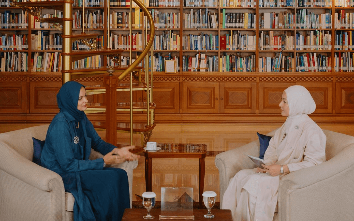 Two women wearing hijabs seated in armchairs having a conversation in a library with a spiral staircase and bookshelves in the background.