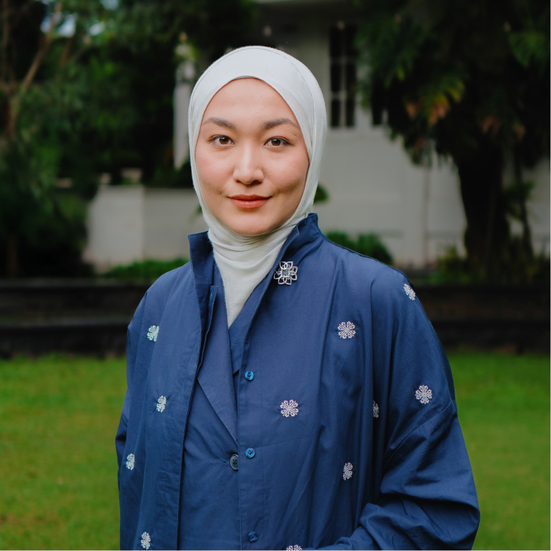 Woman wearing a white hijab and navy blue jacket with floral patterns, standing outdoors with blurred greenery background.