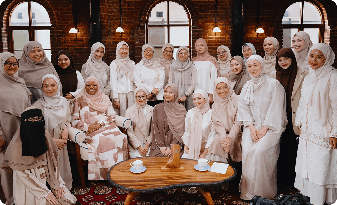 Group of women wearing modest clothing and hijabs, posing in a warmly lit room with brick walls and arched windows behind them.