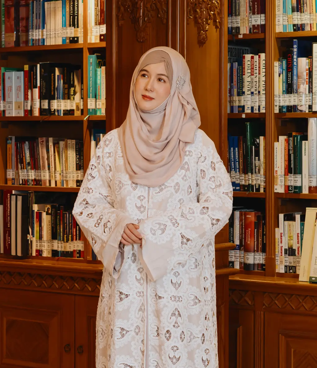 Smiling woman in a white hijab and light blue patterned top sitting on a chair, with another woman in a beige hijab blurred in the background.