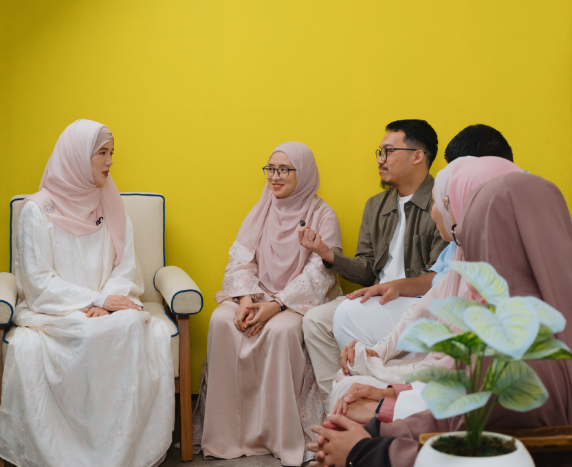 Hand operating a video camera on a tripod capturing a group of women sitting indoors, some wearing hijabs.