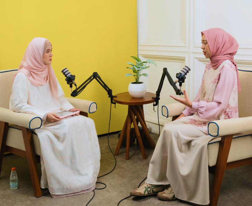 Smiling woman wearing a white hijab and lavender dress, with two other women in hijabs blurred in the background.