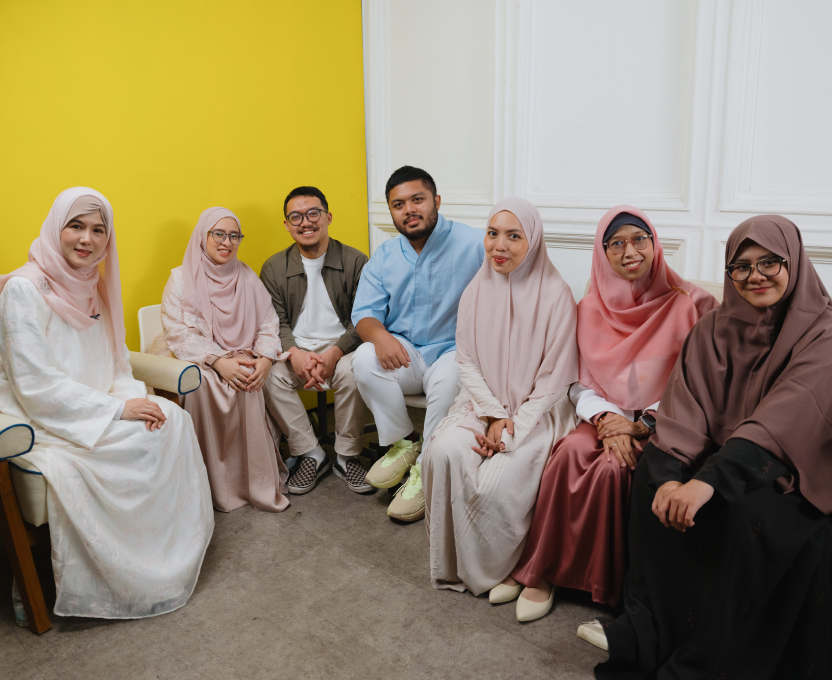 Woman wearing a pale pink hijab speaking in a well-lit room with blurred people in the background.