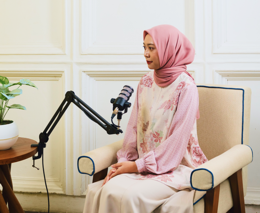 Smiling woman wearing a beige hijab and patterned dress seated, with three women in hijabs in the background.