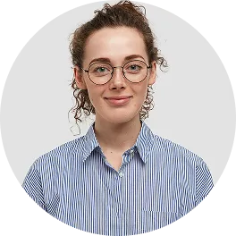 Young woman with curly hair wearing round glasses and a blue striped shirt smiling against a light gray background.