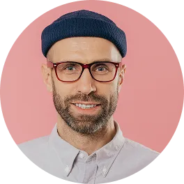 Smiling man wearing a navy beanie, red glasses, and a light gray button-up shirt against a pink background.